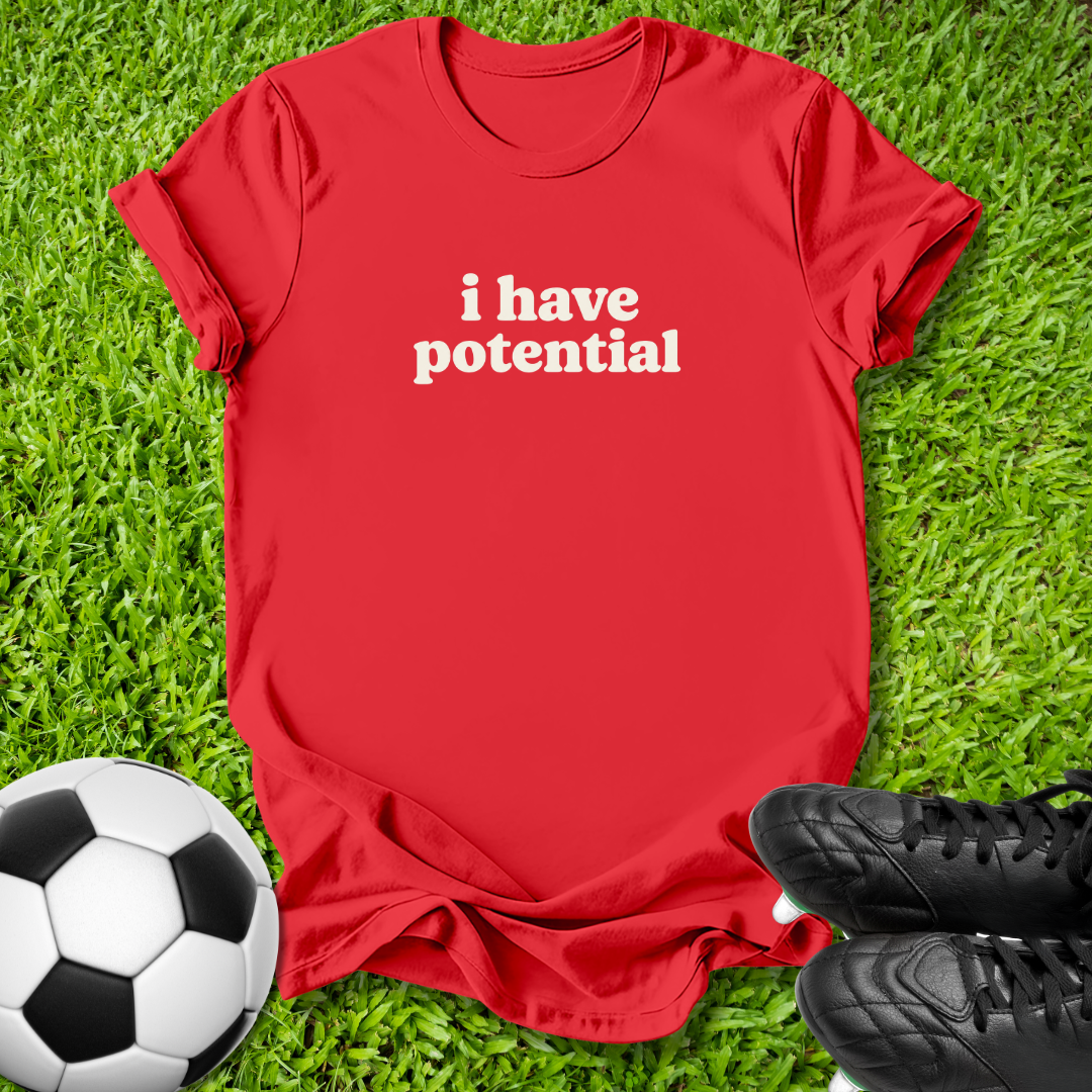 A red short-sleeve t-shirt with the phrase 'i have potential' printed in white lowercase letters across the chest, laid flat on green grass with a soccer ball and black cleats as props.