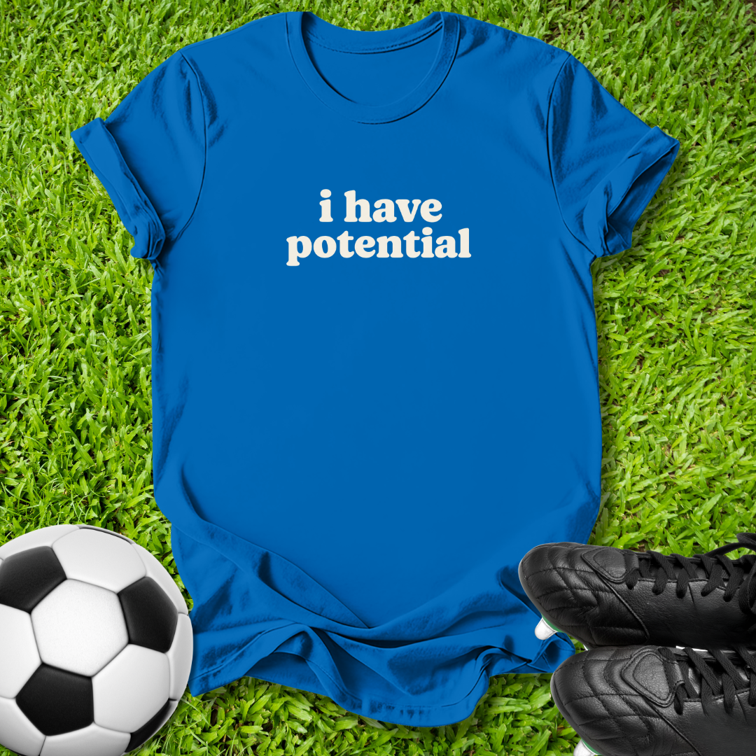 A royal blue short-sleeve t-shirt with the phrase 'i have potential' printed in white lowercase letters across the chest, laid flat on green grass with a soccer ball and black cleats as props.