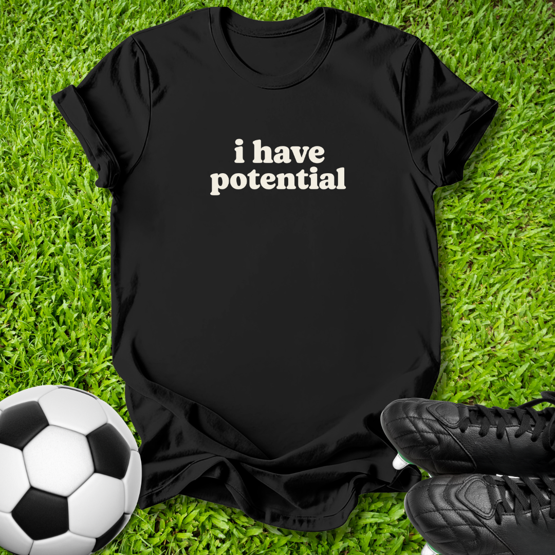 A black short-sleeve t-shirt with the phrase 'i have potential' printed in white text across the chest, laid flat on green grass with a soccer ball and black cleats as props.