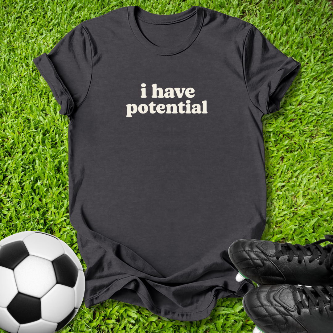 A black short-sleeve t-shirt with the phrase 'i have potential' printed in white lowercase letters across the chest, laid flat on green grass with a soccer ball and black cleats as props.