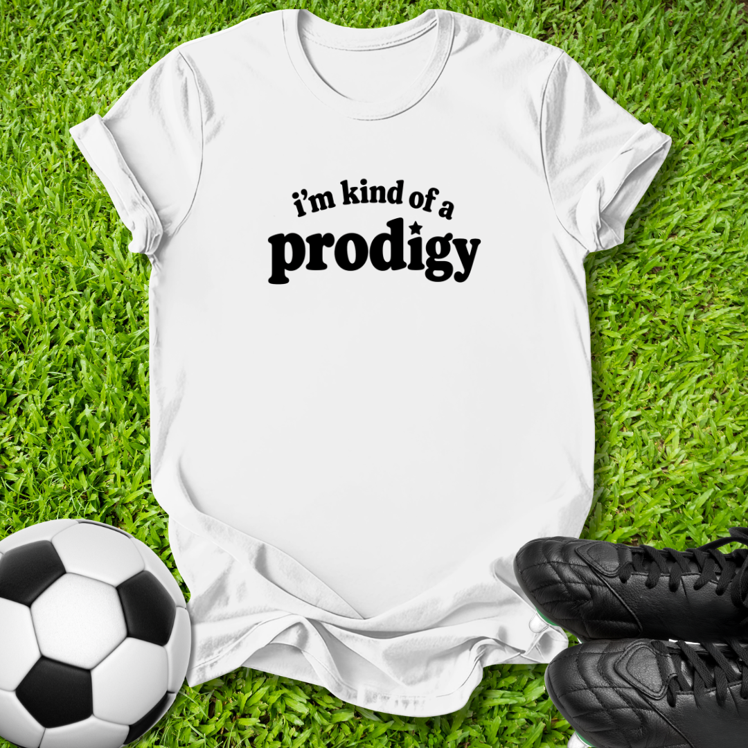 A white short-sleeve t-shirt with the black text 'i'm kind of a prodigy' printed on the chest, laid flat on green grass with a soccer ball and black cleats as props.