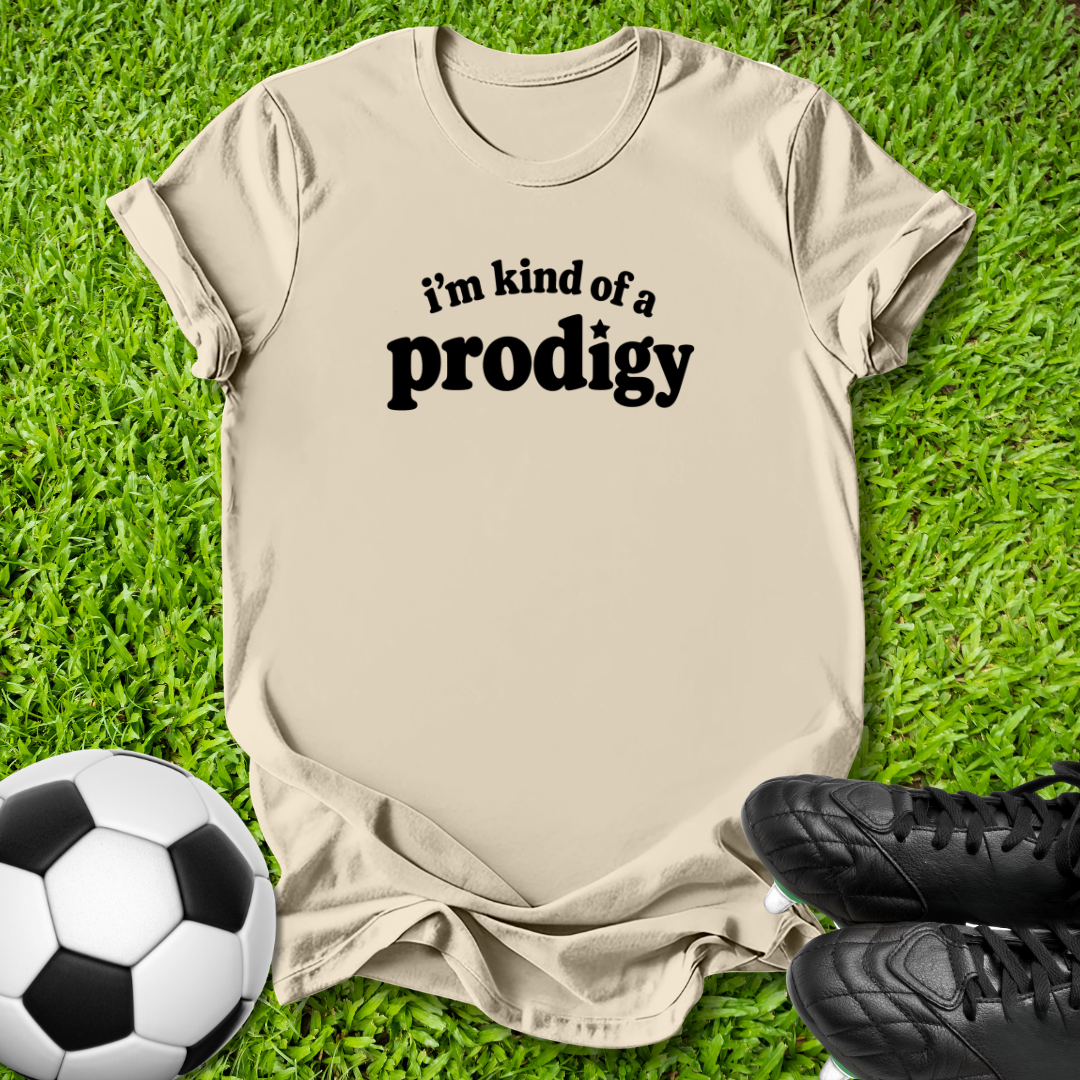 A beige graphic t-shirt with the text 'i'm kind of a prodigy' printed in black, laid flat on green grass with a soccer ball and black cleats as props.