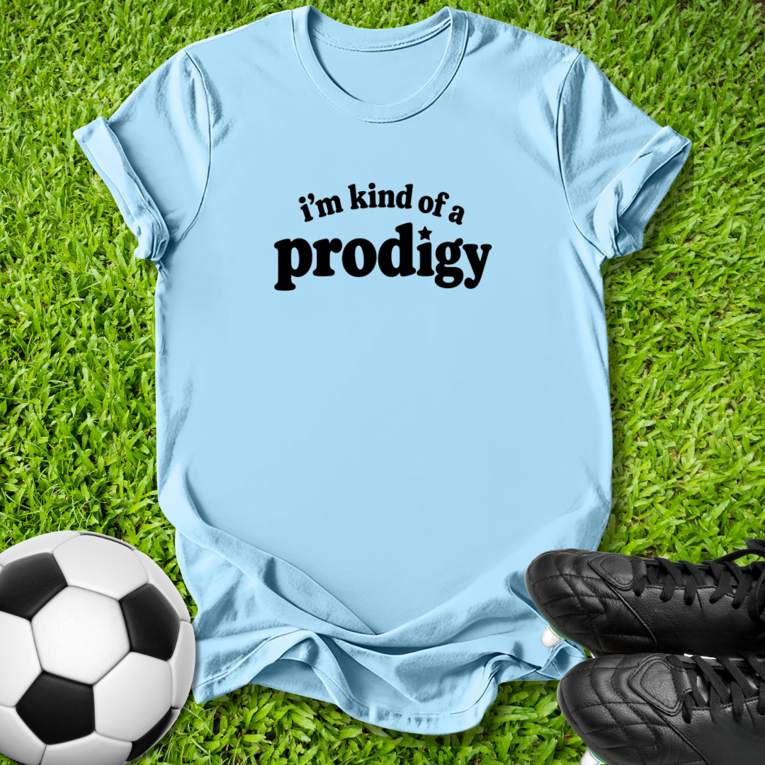 A light blue short-sleeve t-shirt with the black text 'i'm kind of a prodigy' printed on the chest, laid flat on green grass with a soccer ball and black cleats as props.
