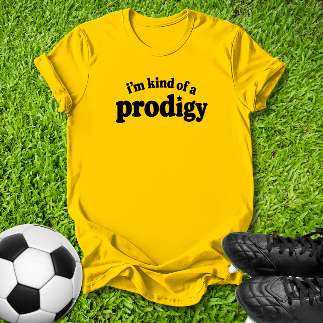 A bright yellow graphic t-shirt laid flat on green grass, featuring the black text 'i'm kind of a prodigy'. A soccer ball is visible in the bottom-left corner and a pair of black soccer cleats is visible in the bottom-right corner.