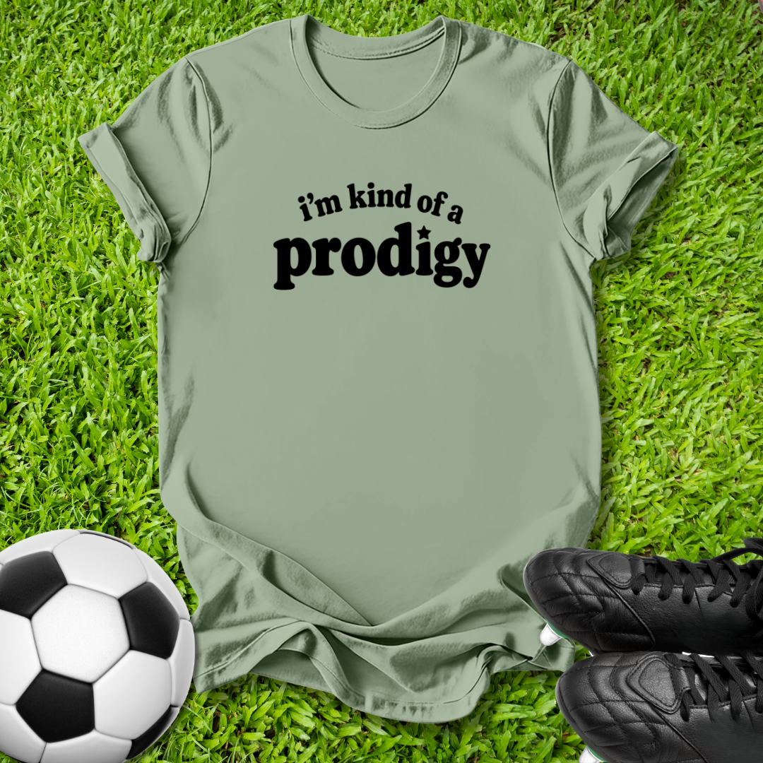 An olive green t-shirt with the black printed text 'i'm kind of a prodigy' lies flat on green grass, styled with a soccer ball on the left and black cleats on the right.