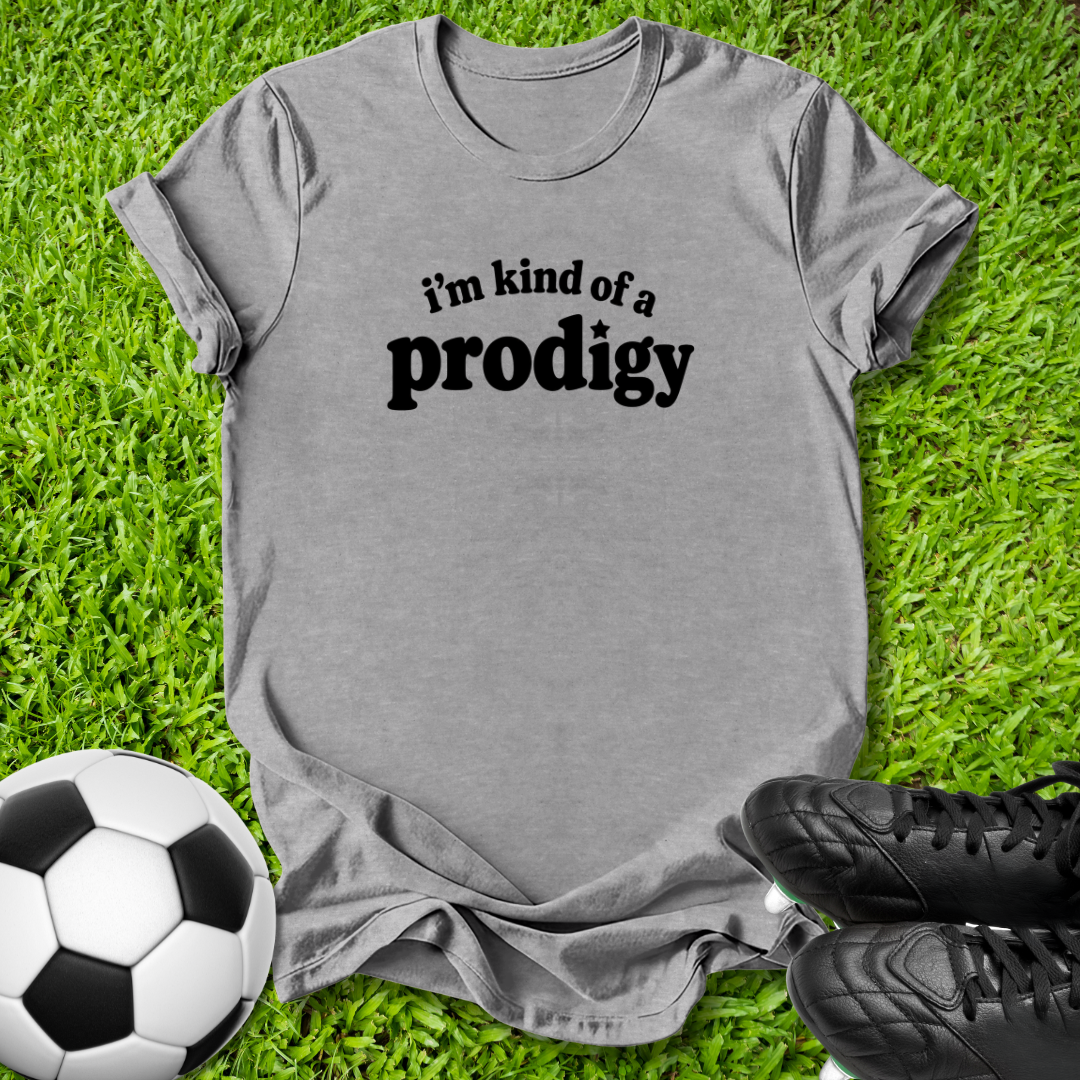 A heather gray short-sleeve t-shirt with the black printed text 'i'm kind of a prodigy' laid flat on green grass, styled with a soccer ball and black cleats.