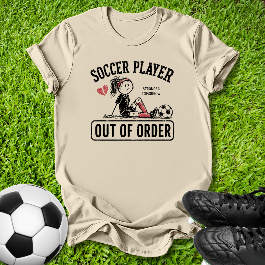 A beige graphic t-shirt laid flat on green grass, featuring a cartoon soccer player illustration with the text 'SOCCER PLAYER OUT OF ORDER'. A soccer ball is on the left and black cleats are on the right.