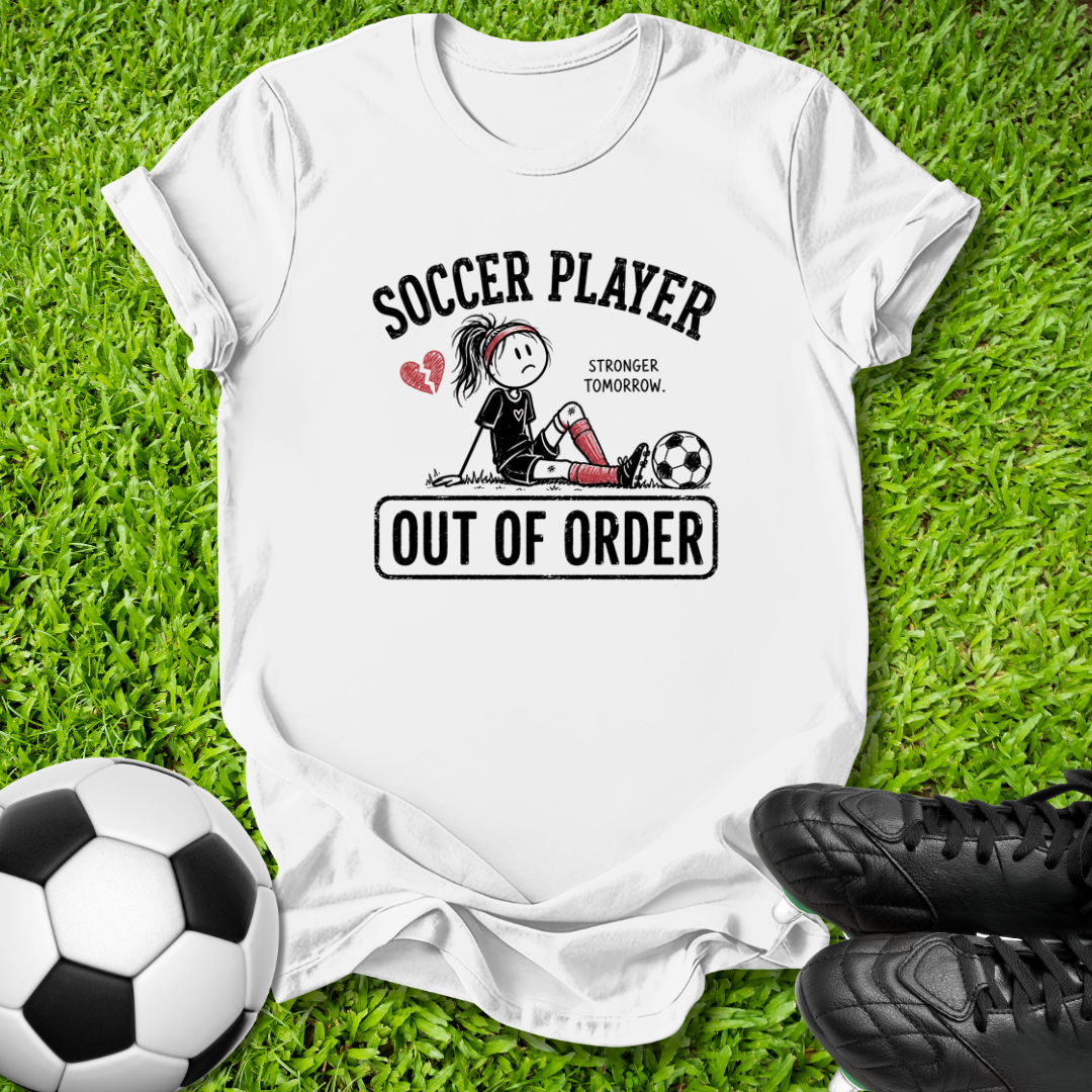 A white graphic t-shirt laid flat on green grass, featuring a soccer-themed design with the text 'SOCCER PLAYER' and 'OUT OF ORDER' along with a cartoon player and soccer ball. A soccer ball and black cleats are placed around it as props.