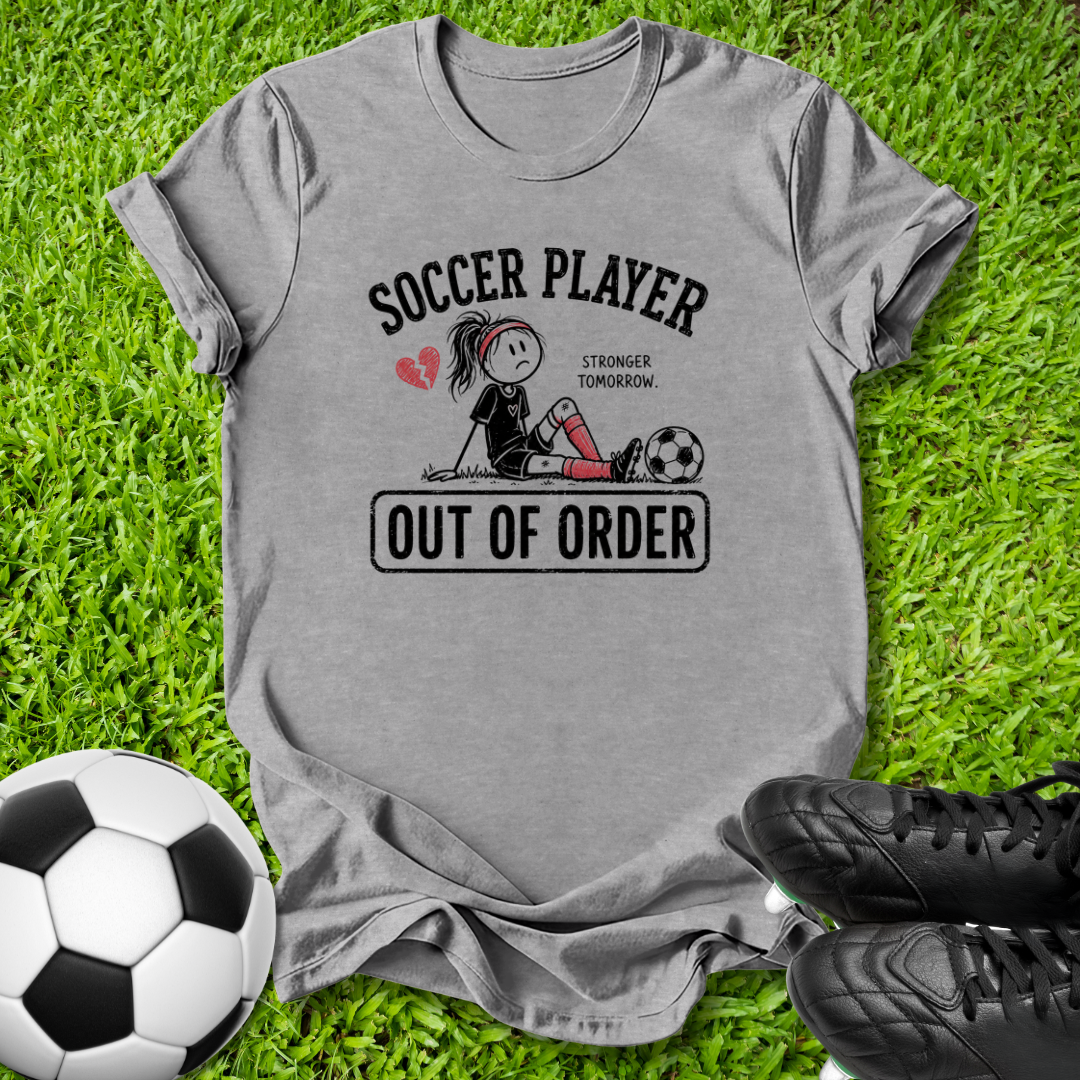 A heather gray graphic t-shirt laid flat on green grass, featuring a soccer player illustration with the text 'SOCCER PLAYER OUT OF ORDER'. A soccer ball is on the bottom left and black cleats are on the bottom right.