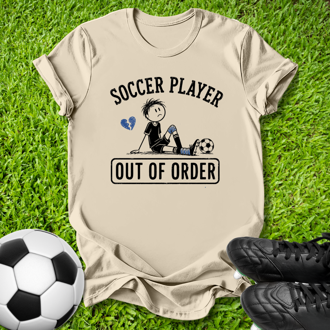 A beige graphic t-shirt laid flat on green grass, featuring a cartoon soccer player design with the text 'SOCCER PLAYER OUT OF ORDER'. A soccer ball and black cleats are placed around it as props.
