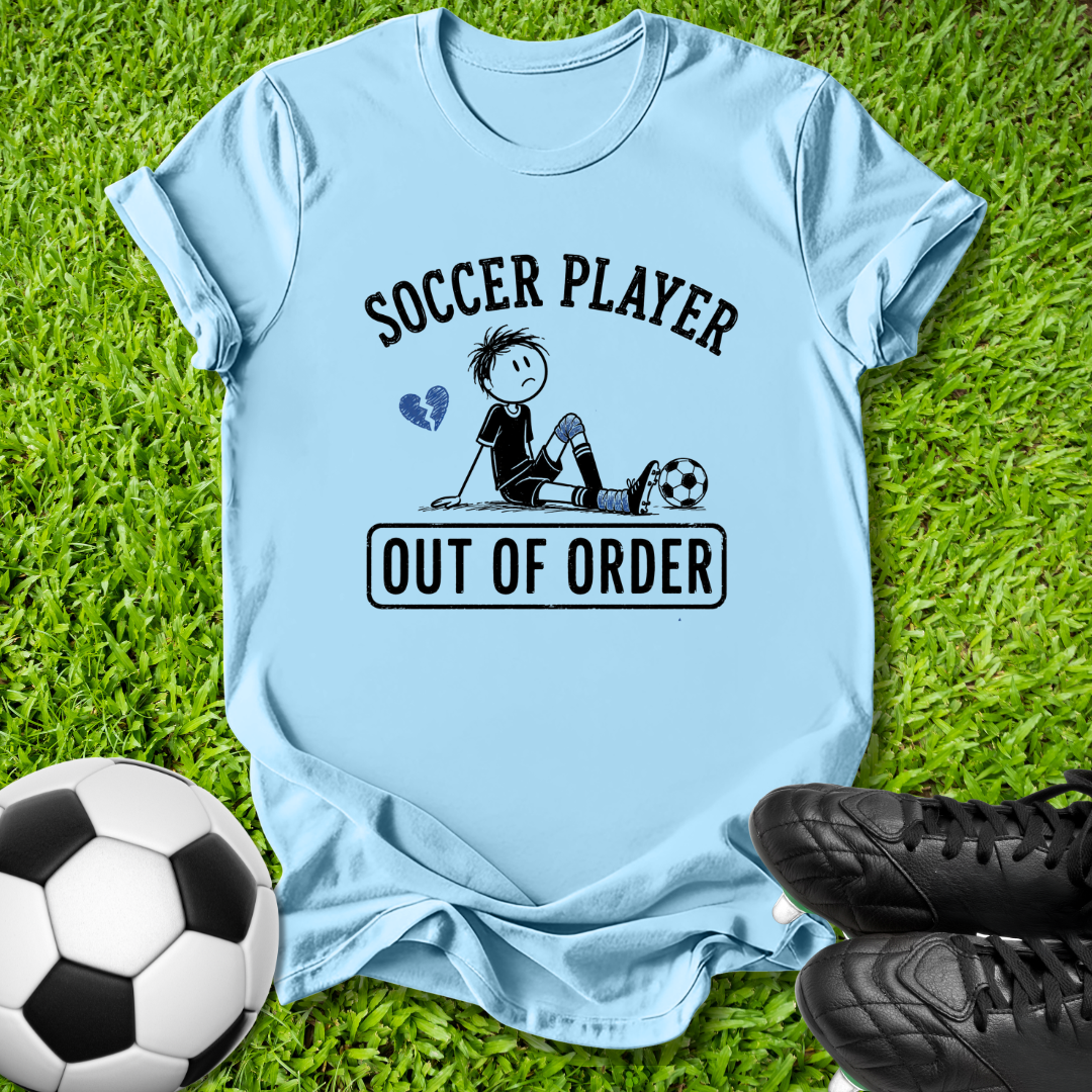 A light blue graphic t-shirt laid flat on green grass, featuring a cartoon soccer player and the text 'SOCCER PLAYER OUT OF ORDER'. A soccer ball and black cleats are placed around it as props.