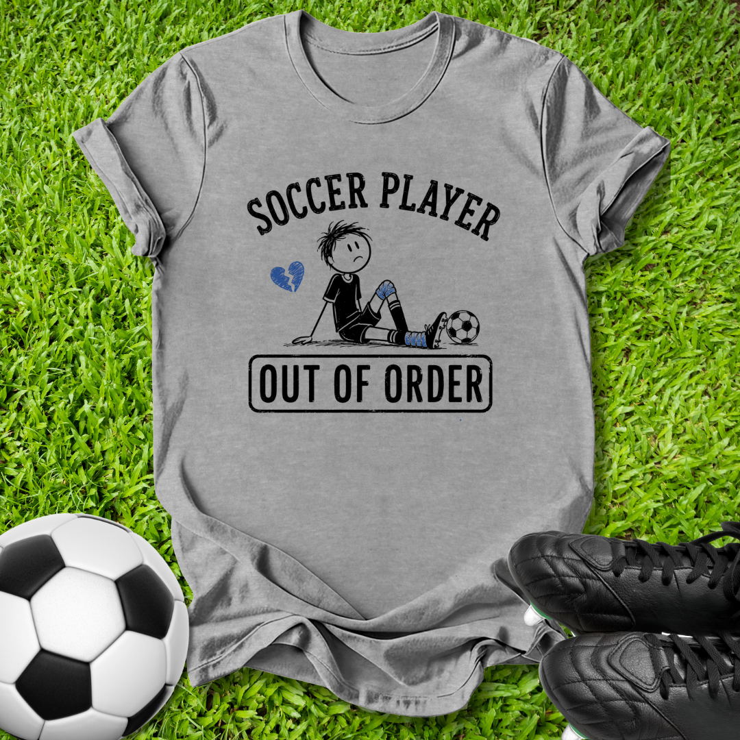 A heather gray graphic t-shirt laid flat on green grass, featuring a cartoon soccer player and the text 'SOCCER PLAYER OUT OF ORDER'. A soccer ball and black cleats are styled around it.