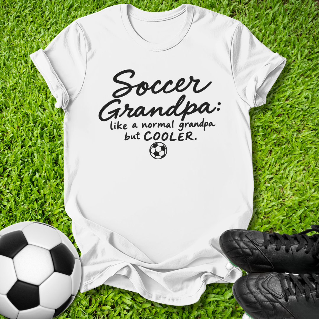 A white short-sleeve graphic T-shirt laid flat on green grass, with a soccer ball at the lower left and black cleats at the lower right. The shirt has black text reading 'Soccer Grandpa: like a normal grandpa but COOLER.