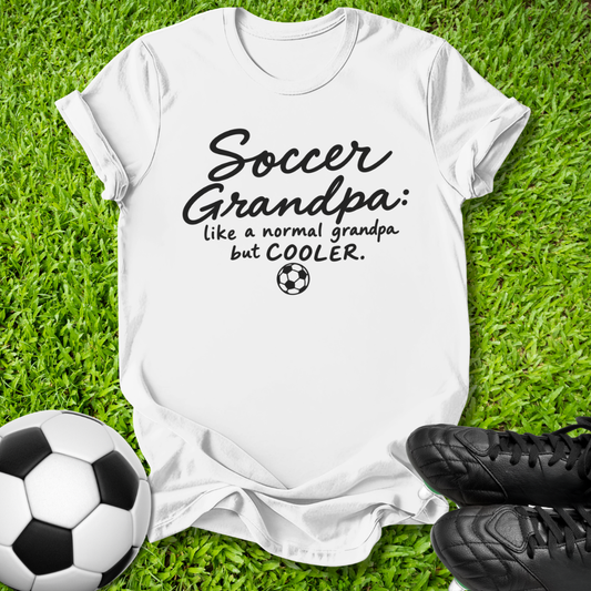 A white short-sleeve graphic T-shirt laid flat on green grass, with a soccer ball at the lower left and black cleats at the lower right. The shirt has black text reading 'Soccer Grandpa: like a normal grandpa but COOLER.