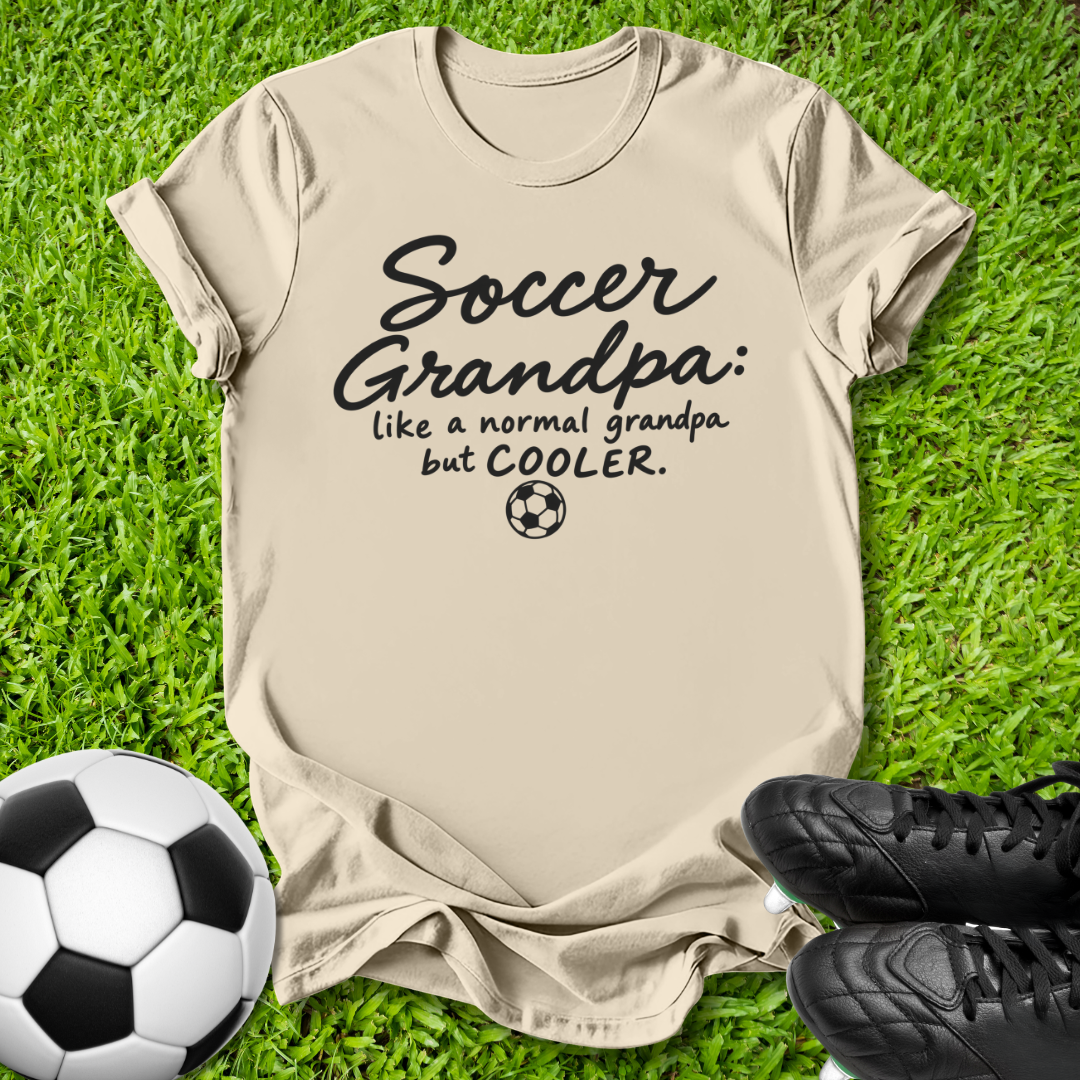 A beige graphic t-shirt laid flat on green grass, featuring the text 'Soccer Grandpa: like a normal grandpa but COOLER.' with a small soccer ball icon.
