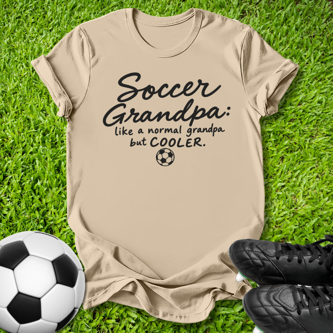 A beige graphic t-shirt laid flat on green grass, featuring the text 'Soccer Grandpa: like a normal grandpa but COOLER.' in black script and print with a small soccer ball icon.
