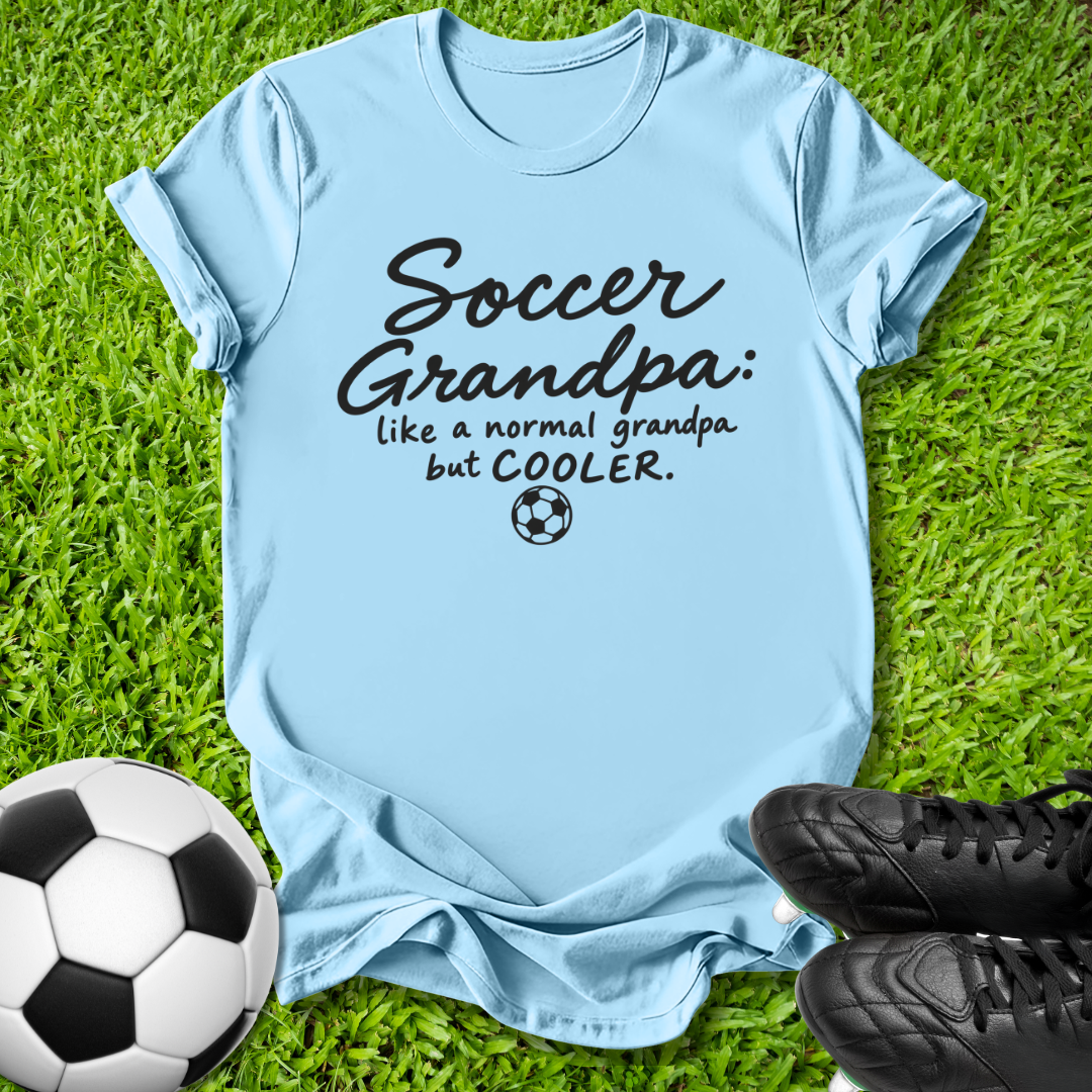 A light blue graphic t-shirt laid flat on green grass, featuring the text 'Soccer Grandpa: like a normal grandpa but COOLER.' in black script and print with a small soccer ball icon.