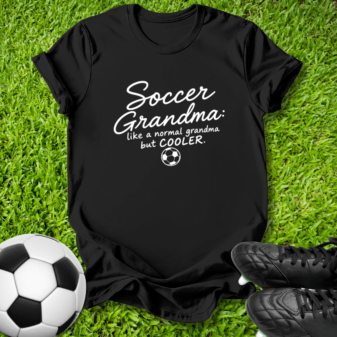 A black short-sleeve graphic T-shirt laid flat on green grass, with a soccer ball at the lower left and black cleats at the lower right. The shirt has white text reading 'Soccer Grandma like a normal grandma but COOLER' with a small soccer ball graphic.