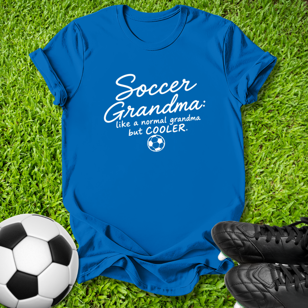 A royal blue short-sleeve t-shirt laid flat on green grass, featuring the white printed text 'Soccer Grandma like a normal grandma but COOLER.' with a small soccer ball graphic.