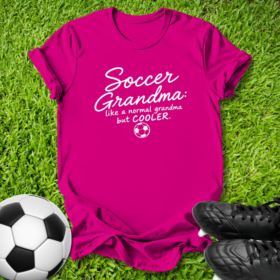 A bright pink short-sleeve t-shirt laid flat on green grass, featuring the white printed text 'Soccer Grandma like a normal grandma but COOLER' with a small soccer ball graphic. A black-and-white soccer ball is at the lower left, and black cleats are at the lower right.