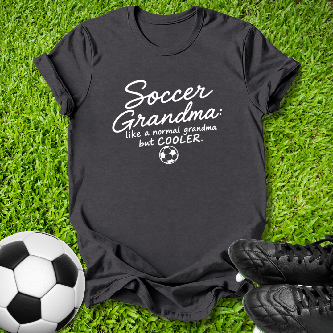A black graphic t-shirt laid flat on green grass with a soccer ball and black cleats nearby. The shirt features the text 'Soccer Grandma like a normal grandma but COOLER.