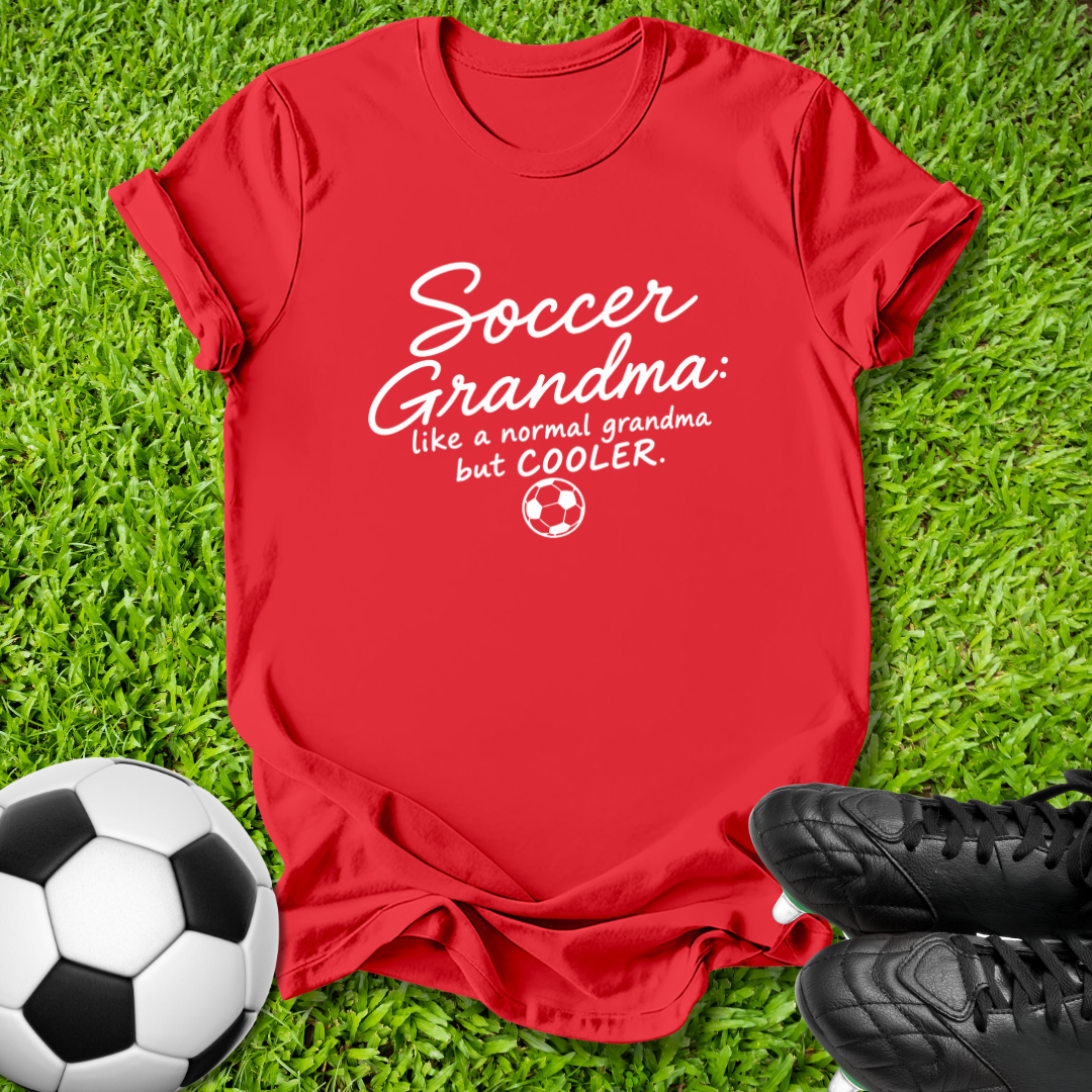 A red short-sleeve graphic T-shirt laid flat on green grass, with a soccer ball at the lower left and black cleats at the lower right. The shirt has white text reading 'Soccer Grandma like a normal grandma but COOLER' with a small soccer ball graphic.