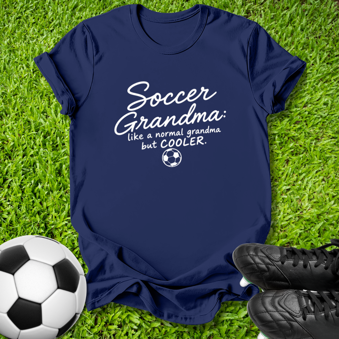 A navy blue t-shirt with a white graphic reading 'Soccer Grandma like a normal grandma but COOLER.' with a small soccer ball icon, laid flat on green grass with a soccer ball and black cleats nearby.