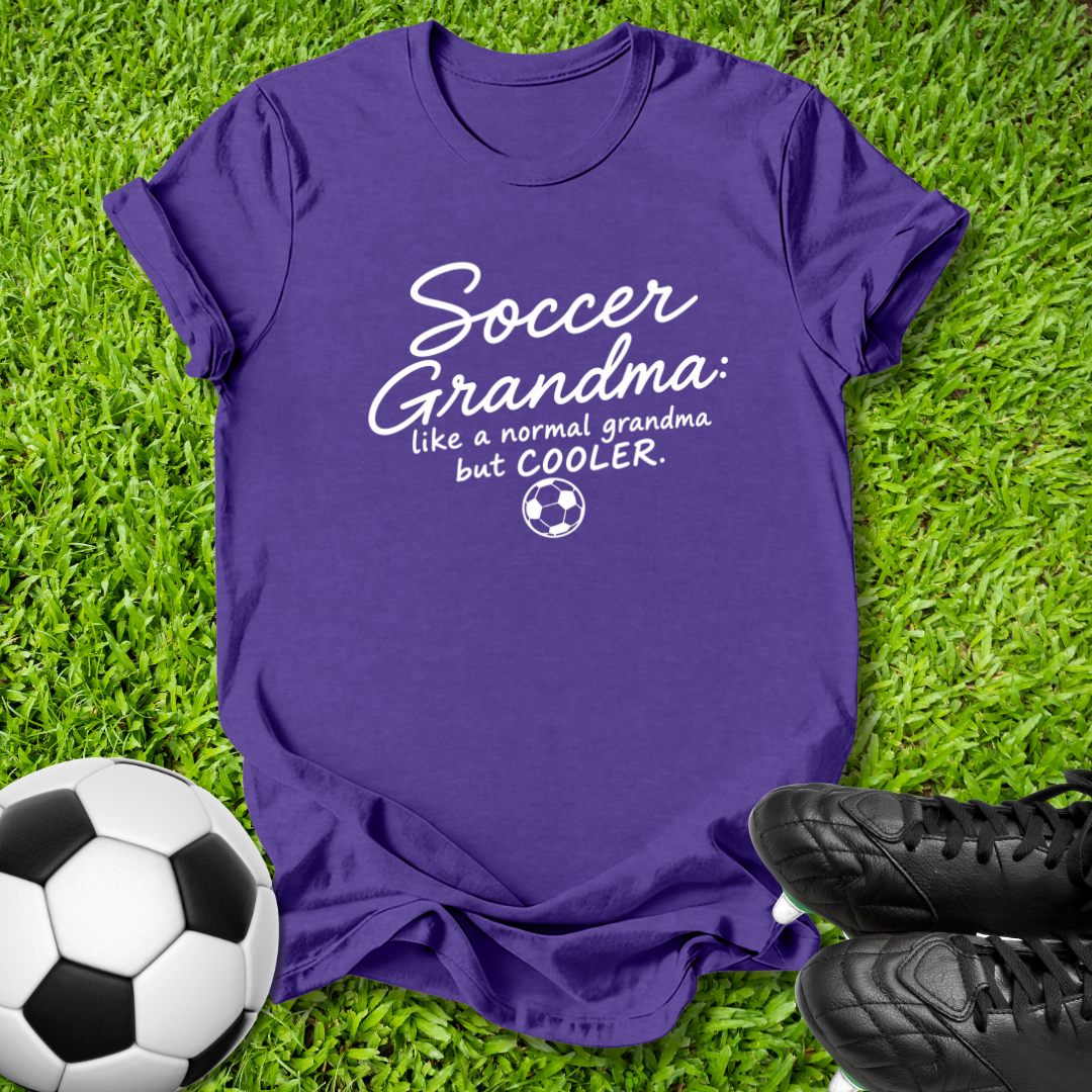 A purple short-sleeve t-shirt with a white graphic reading 'Soccer Grandma like a normal grandma but COOLER' with a small soccer ball icon, laid flat on green grass with a soccer ball and black cleats nearby.