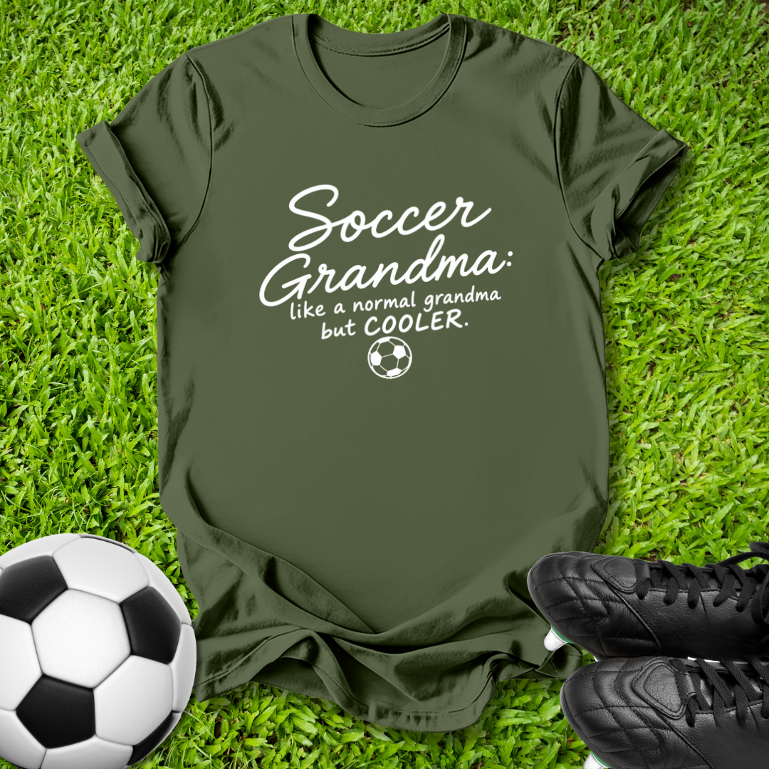 An olive green short-sleeve t-shirt with a white graphic text reading 'Soccer Grandma like a normal grandma but COOLER.' with a small soccer ball icon, laid flat on green grass with a soccer ball and black cleats as props.