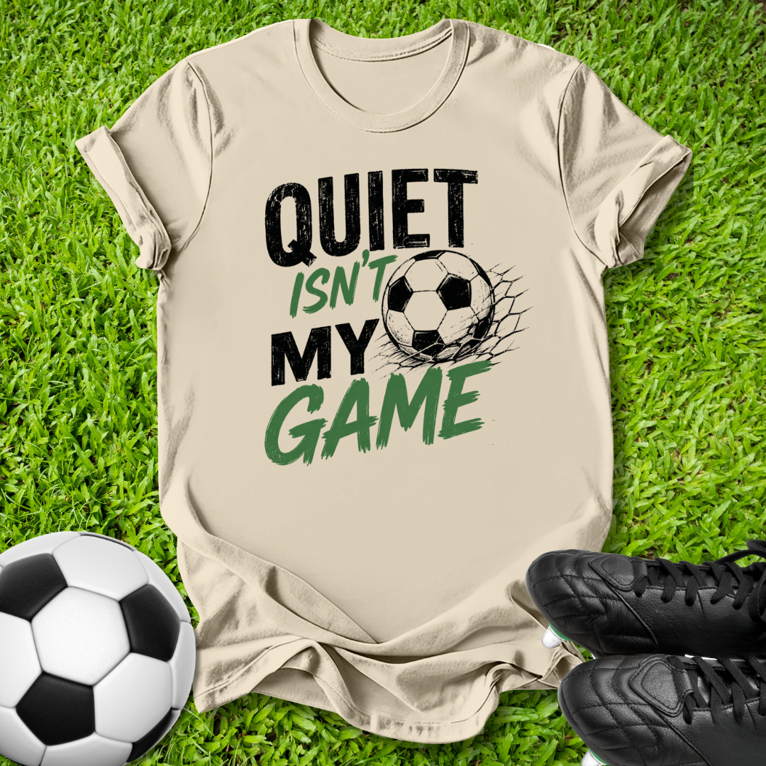 A beige graphic t-shirt laid flat on green grass, featuring the text 'QUIET ISN'T MY GAME' in black and green with a soccer ball design. A soccer ball and black cleats are placed around it as props.