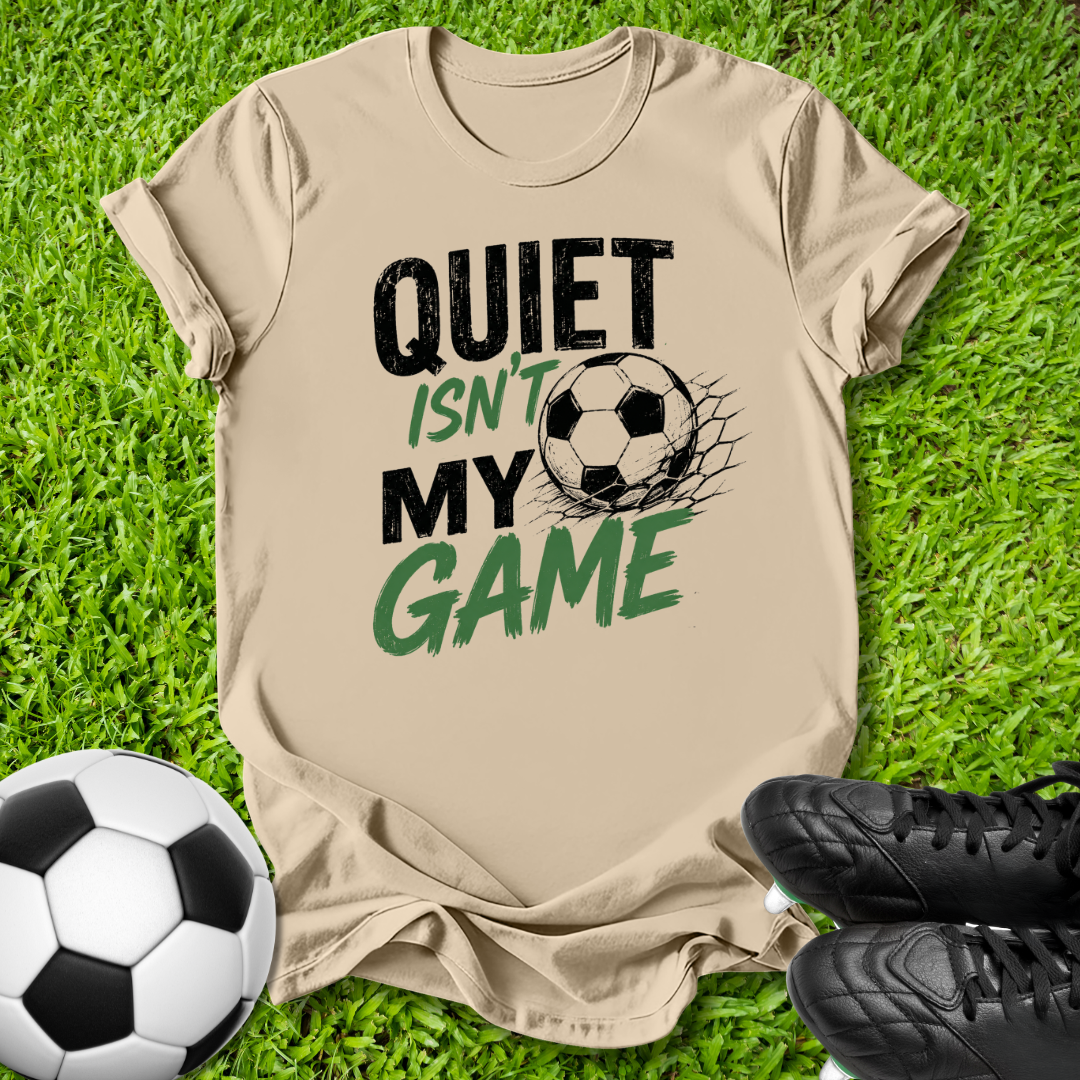 A beige graphic t-shirt laid flat on green grass, featuring the text 'QUIET ISN'T MY GAME' in black and green with a soccer ball graphic. A real soccer ball and black cleats are placed around it as props.