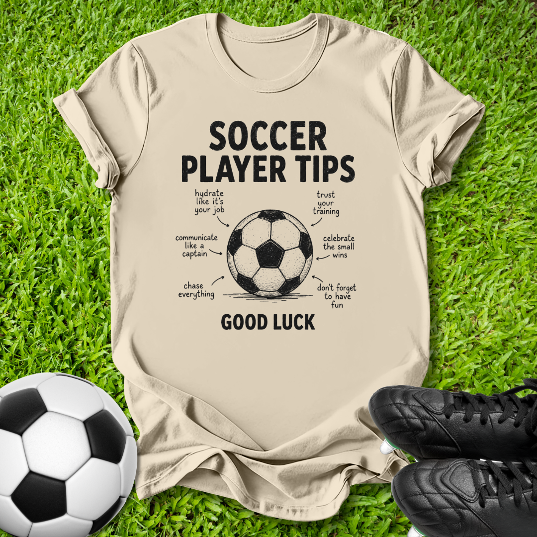 A beige graphic t-shirt laid flat on green grass, featuring a soccer theme with the text 'SOCCER PLAYER TIPS' and 'GOOD LUCK' around a soccer ball illustration. A real soccer ball and black cleats are placed as props around the shirt.