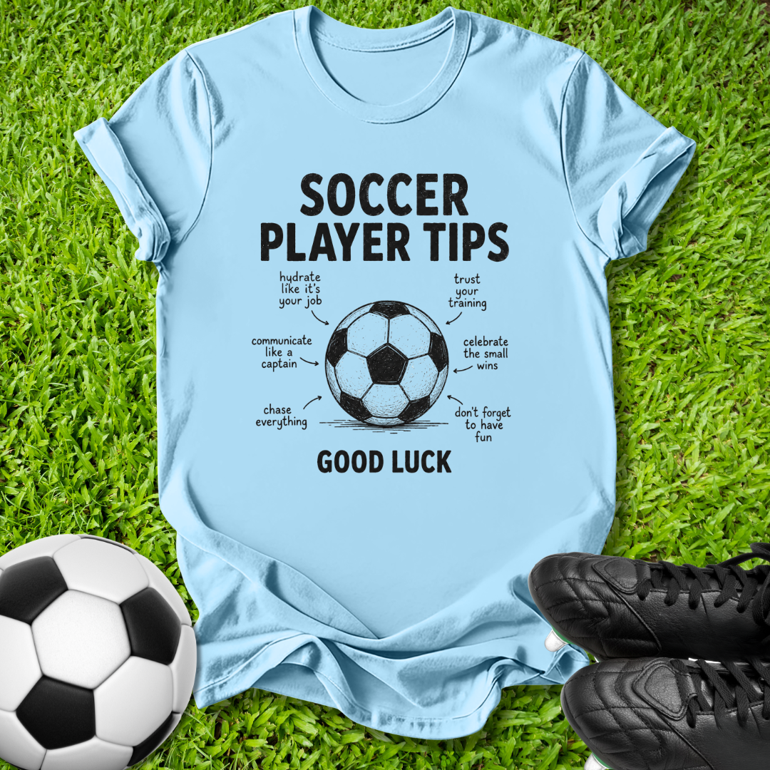 A light blue graphic t-shirt laid flat on green grass, featuring a soccer ball illustration and the text 'SOCCER PLAYER TIPS' and 'GOOD LUCK' with handwritten-style notes around it. A black-and-white soccer ball sits at the lower left, and black cleats are at the lower right.