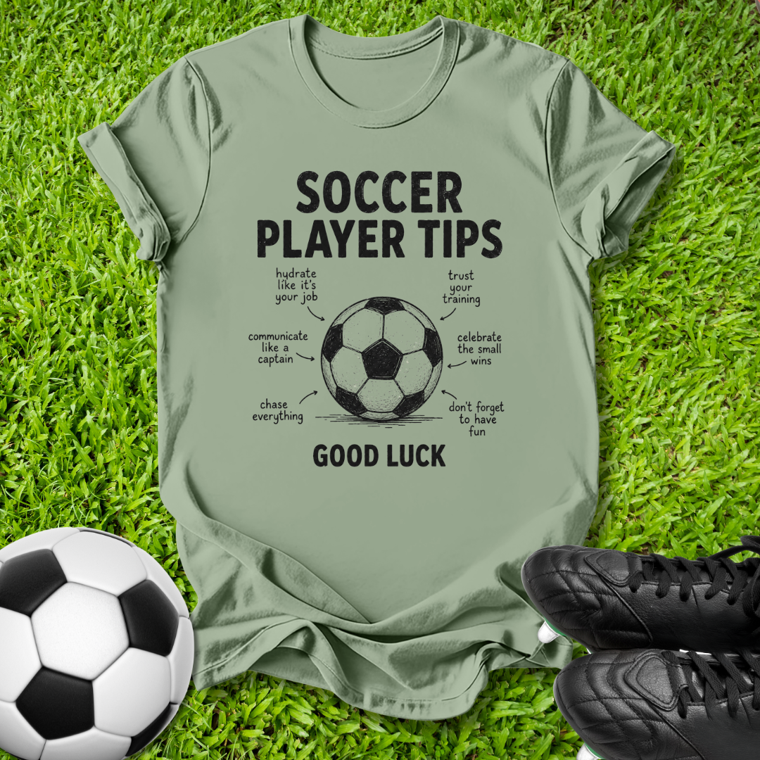 An olive green graphic t-shirt laid flat on green grass, featuring a black and white soccer ball illustration with the text 'SOCCER PLAYER TIPS' and 'GOOD LUCK' surrounding it. A real soccer ball and black cleats are placed as props around the shirt.