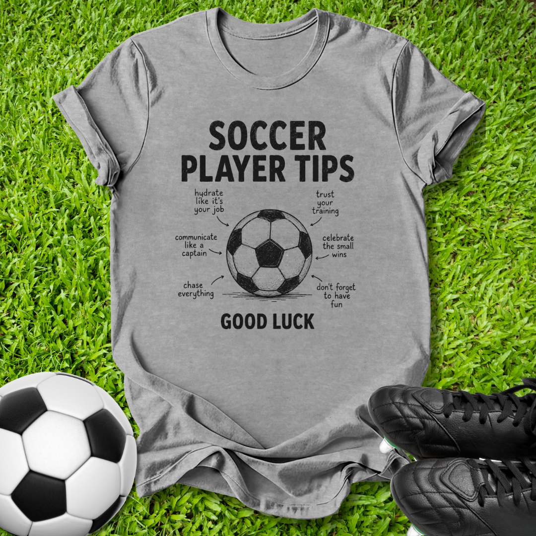 A heather gray short-sleeve t-shirt with a black graphic print reading 'SOCCER PLAYER TIPS' and 'GOOD LUCK' around a soccer ball illustration, laid flat on green grass with a soccer ball and black cleats nearby.