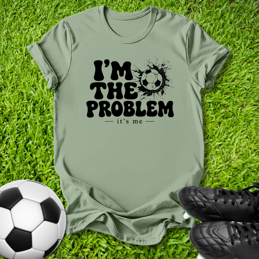 An olive green t-shirt with a black graphic reading 'I'M THE PROBLEM - it's me' next to a soccer ball and soccer cleats, laid flat on green grass.