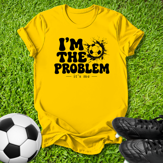 A bright yellow graphic t-shirt laid flat on green grass, featuring the black text 'I'M THE PROBLEM it's me' with a soccer ball splatter design. A soccer ball and black cleats are placed around it as props.