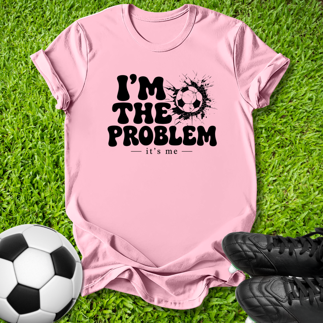 A pink short-sleeve t-shirt with a black graphic reading 'I'M THE PROBLEM it's me' and a soccer ball design, laid flat on green grass with a soccer ball and black cleats nearby.