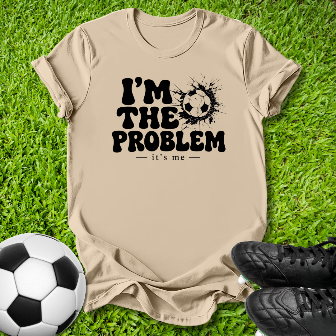 A beige graphic t-shirt laid flat on green grass, featuring the black text 'I'M THE PROBLEM - it's me' with a soccer ball splatter design. A soccer ball and black cleats are styled as props around the shirt.
