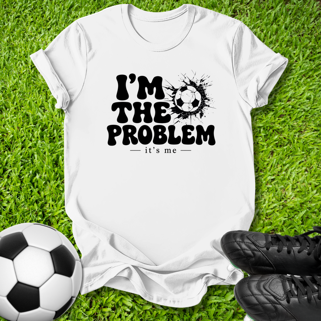 A white short-sleeve graphic T-shirt laid flat on green grass, with a black printed slogan reading “I’M THE PROBLEM it’s me” alongside a soccer ball splatter graphic. A real soccer ball and black cleats are placed around the shirt as props.