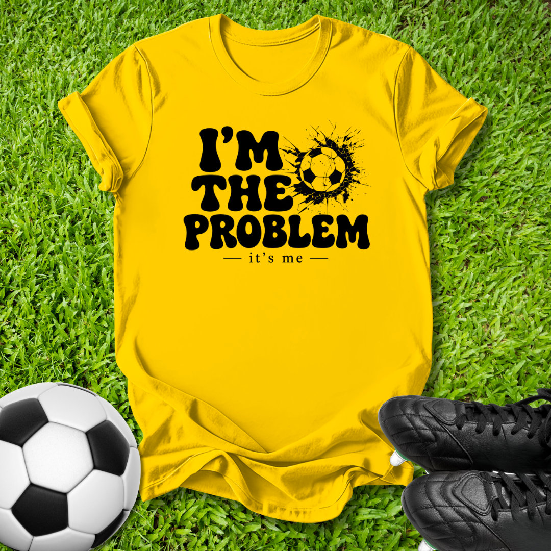 A bright yellow graphic t-shirt laid flat on green grass, featuring the black text 'I'M THE PROBLEM it's me' with a soccer ball splatter design. A soccer ball and black cleats are placed around it as props.
