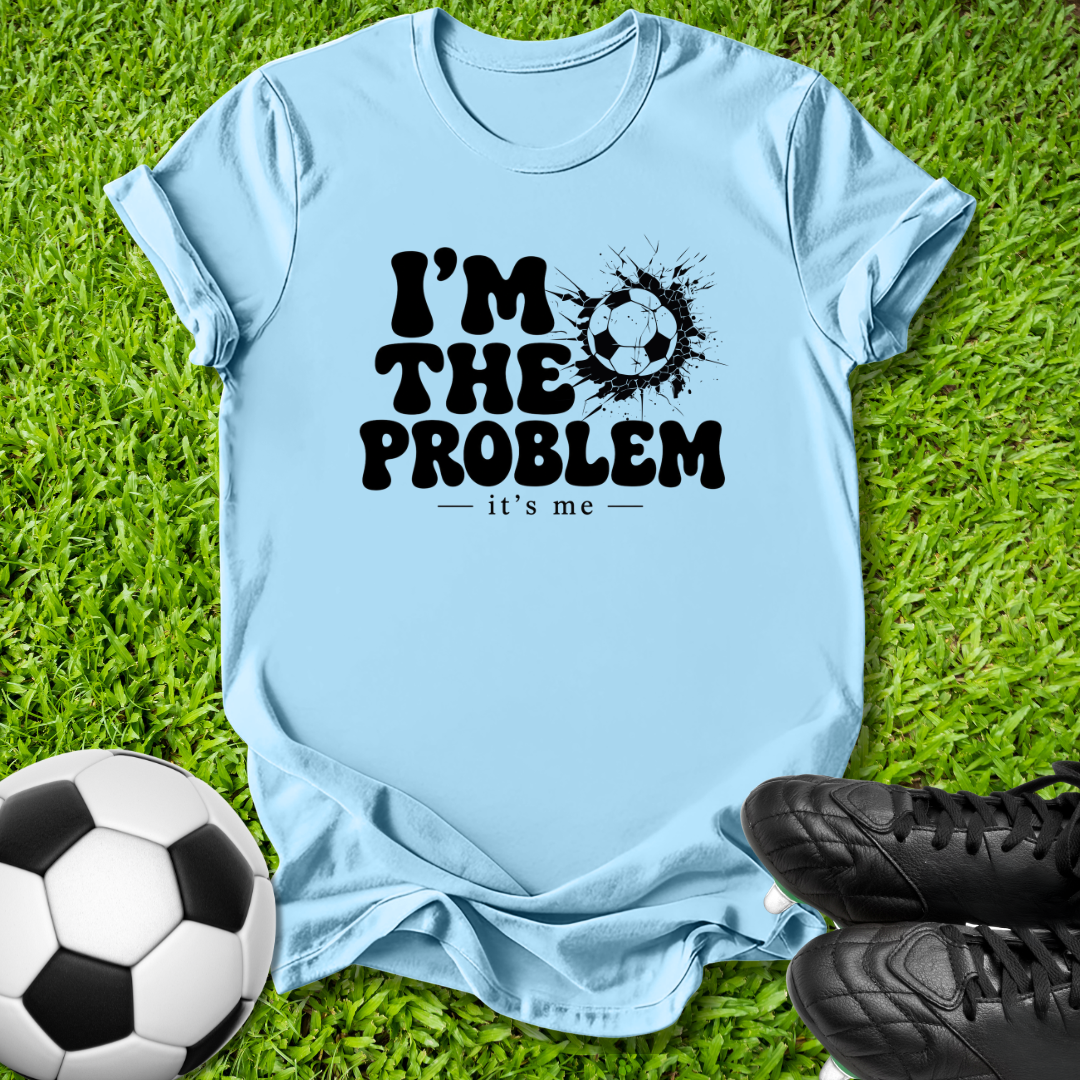 A light blue graphic t-shirt laid flat on green grass, featuring the black text 'I'M THE PROBLEM - it's me' with a soccer ball splatter design. A soccer ball and black cleats are placed around it as props.