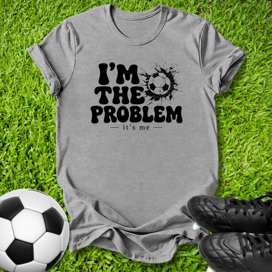 A heather gray short-sleeve t-shirt with a black graphic reading 'I'M THE PROBLEM - it's me' next to a soccer ball, laid flat on green grass with soccer cleats nearby.