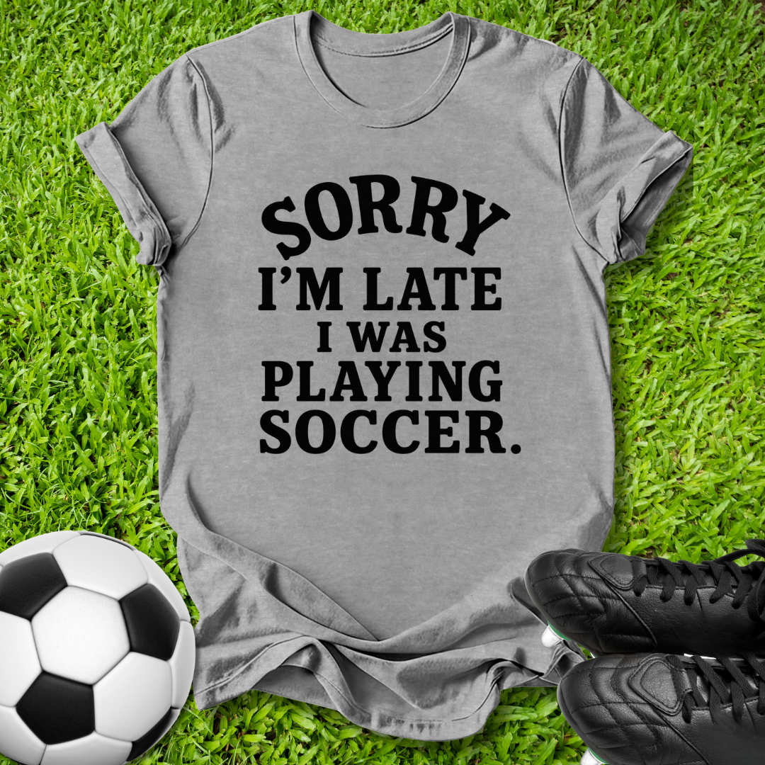 A heather gray short-sleeve t-shirt with black text reading 'SORRY I'M LATE I WAS PLAYING SOCCER.' laid flat on green grass, styled with a soccer ball and black cleats.