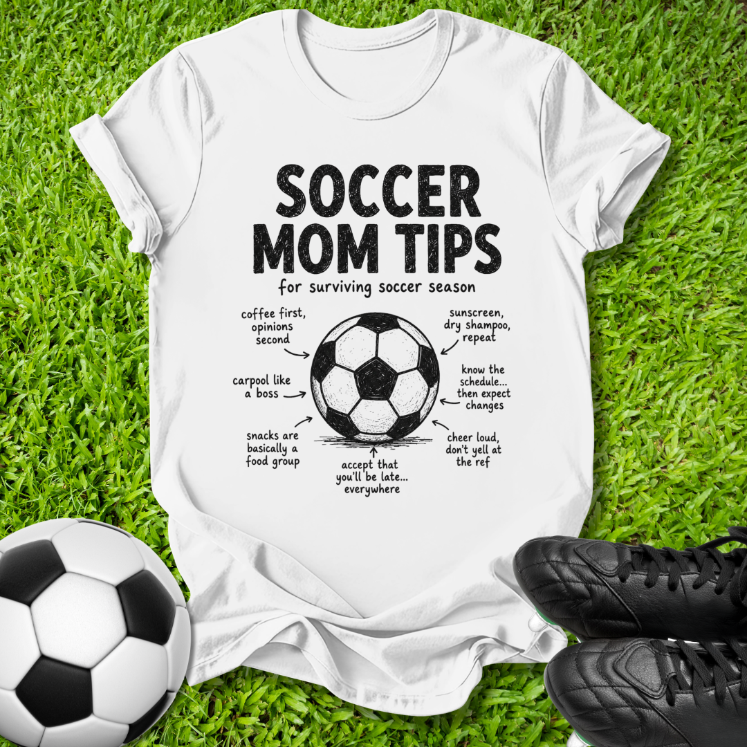 A white graphic t-shirt laid flat on green grass, featuring a black 'Soccer Mom Tips' diagram with a soccer ball illustration and handwritten-style notes. A soccer ball is at the bottom left, and black cleats are at the bottom right.