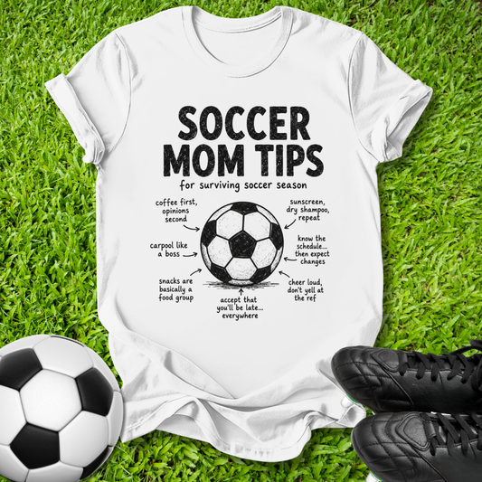 A white graphic t-shirt laid flat on green grass, featuring a black 'Soccer Mom Tips' diagram with a soccer ball illustration and handwritten-style notes. A soccer ball is at the bottom left, and black cleats are at the bottom right.