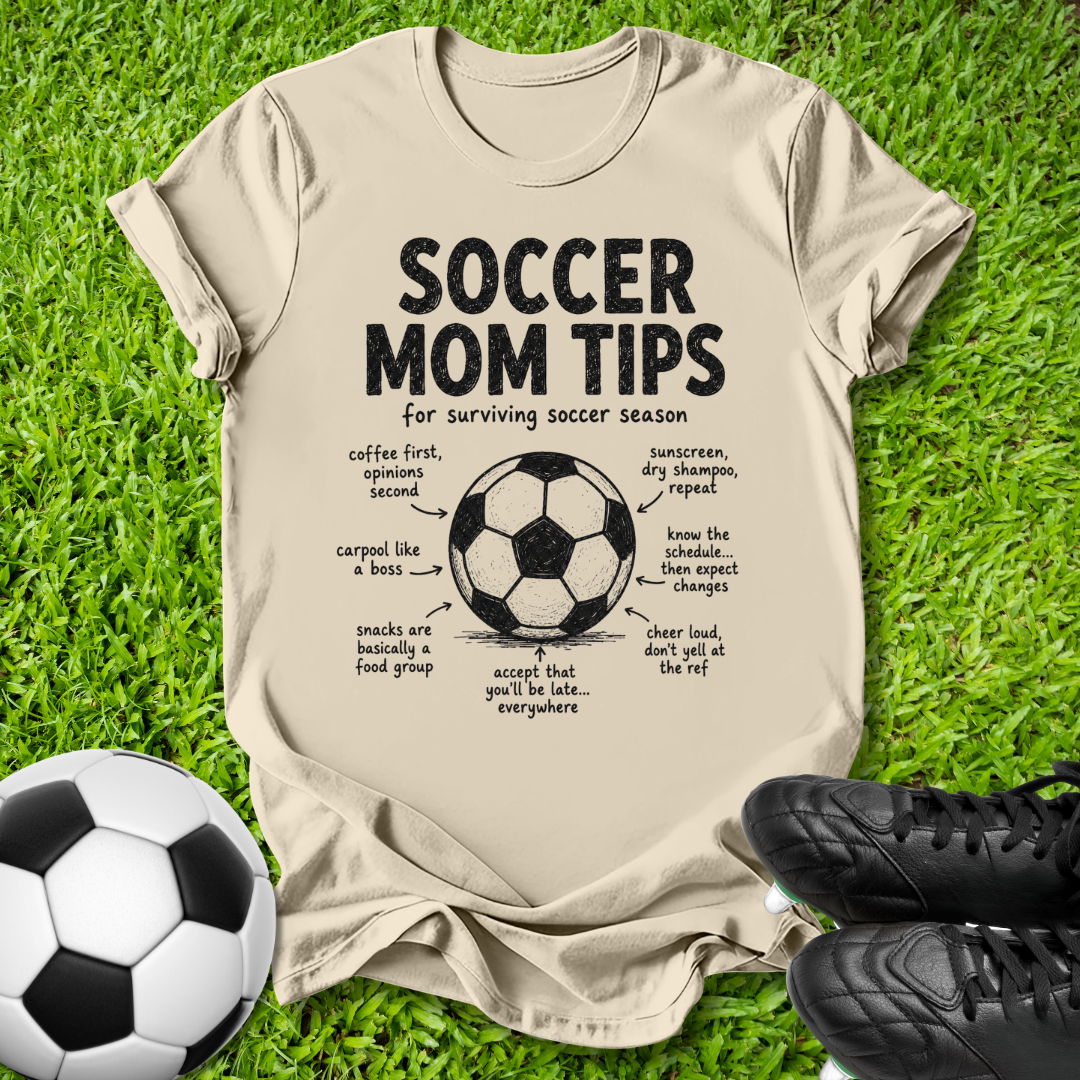 A beige graphic t-shirt laid flat on green grass, featuring a soccer-themed 'Soccer Mom Tips' print with a soccer ball illustration and handwritten-style notes. A black-and-white soccer ball sits at the lower left, and black cleats are at the lower right.