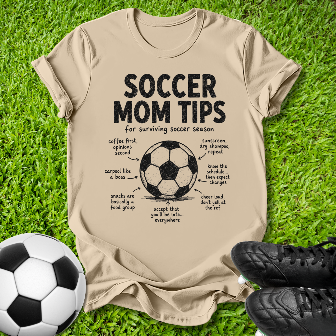 A beige graphic t-shirt laid flat on green grass, featuring a black 'Soccer Mom Tips' design with a soccer ball diagram and handwritten notes. A black-and-white soccer ball sits at the bottom left, and black cleats are at the bottom right.