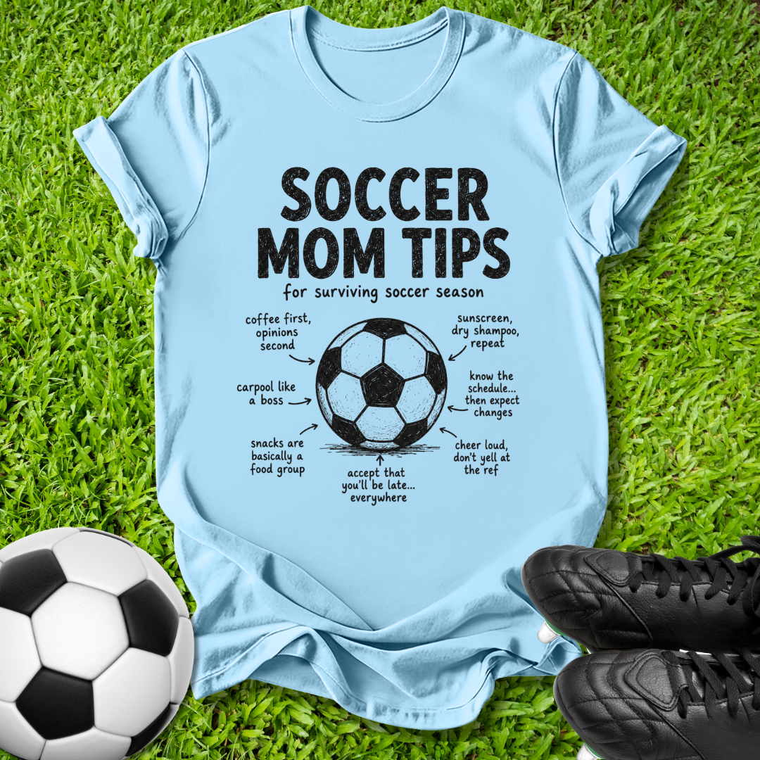 A light blue graphic t-shirt laid flat on green grass, featuring a black 'SOCCER MOM TIPS' design with a soccer ball diagram and handwritten notes. A black-and-white soccer ball sits at the bottom left, and black cleats are at the bottom right.