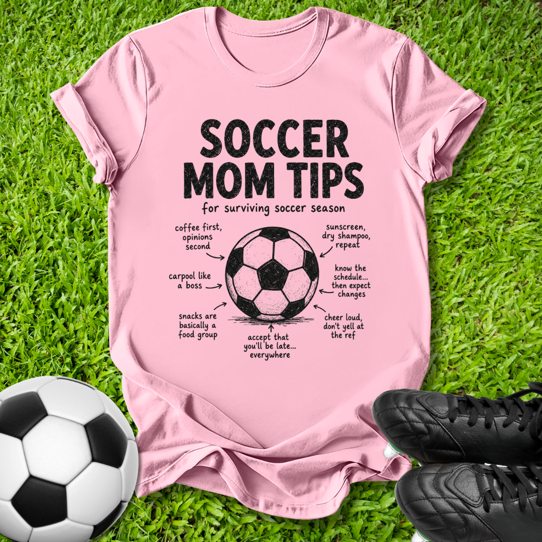 A pink short-sleeve graphic T-shirt laid flat on green grass, featuring a soccer ball illustration and the text 'SOCCER MOM TIPS for surviving soccer season' with handwritten-style notes around it. A soccer ball and black cleats are placed around the shirt as props.
